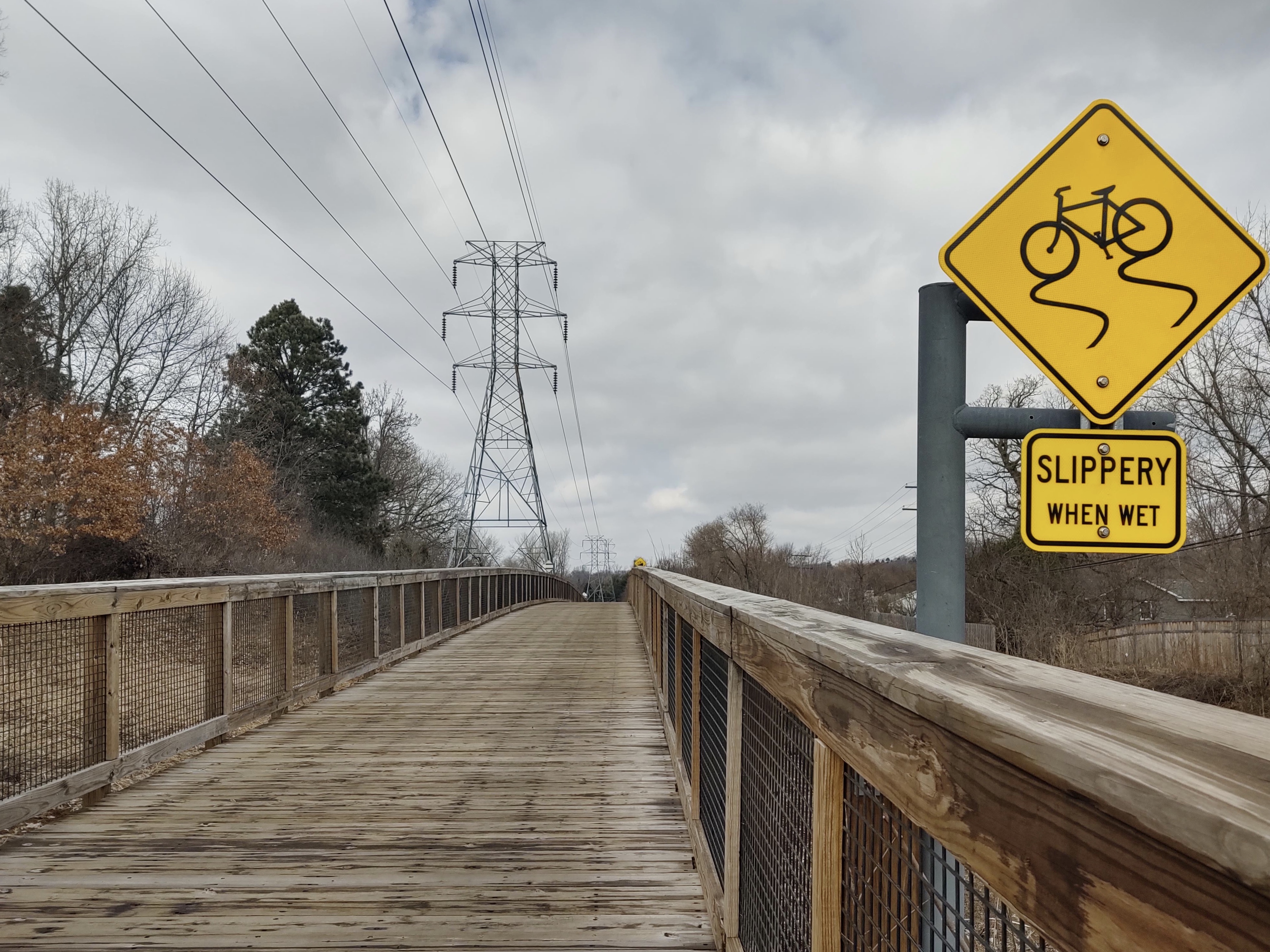 Wooden trail boardwalk beneath utility lines with a bicycle caution sign.